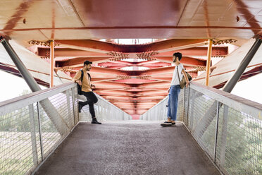 Young gay couple standing on modern footbridge looking at each other - FBAF00069