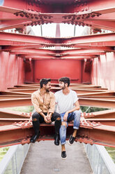 Young gay couple in love sitting on steel girders of a footbridge looking at each other - FBAF00076