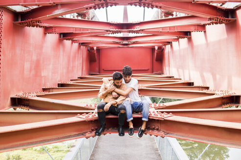 Young gay couple in love sitting on steel girder of a footbridge looking down - FBAF00079