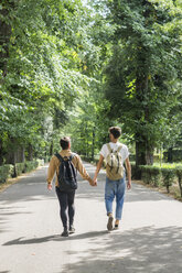 Back view of young gay couple with backpacks walking hand in hand on a road - FBAF00083