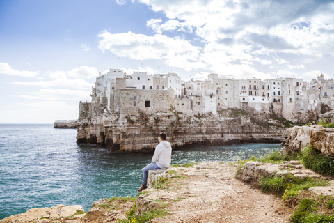 Italien, Apulien, Polognano a Mare, Rückenansicht eines Mannes, der sich auf einem Felsen ausruht und auf den Horizont schaut, lizenzfreies Stockfoto