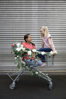 Zwei lächelnde Mädchen, die in einem mit Kunstblumen geschmückten Einkaufswagen hocken - PSTF00175