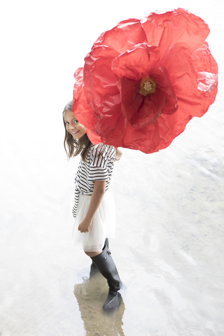 Porträt eines lächelnden Mädchens, das mit einer übergroßen roten Kunstblume in einem See steht, lizenzfreies Stockfoto