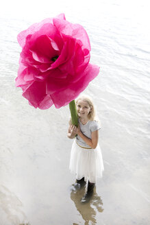 Porträt eines lächelnden blonden Mädchens, das in einem See steht und eine übergroße rosa Kunstblume hält - PSTF00212