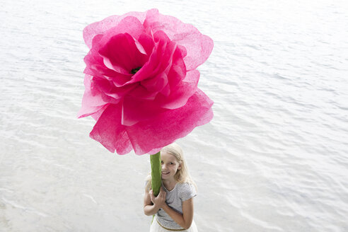 Porträt eines lächelnden blonden Mädchens, das vor einem See steht und eine übergroße rosa Kunstblume hält - PSTF00213