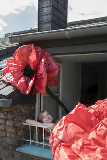 Glückliches Mädchen mit übergroßer roter Kunstblume, das sich aus dem Fenster lehnt - PSTF00245