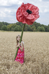 Porträt eines lächelnden Mädchens, das auf einem Feld mit einer übergroßen roten Kunstblume steht - PSTF00250