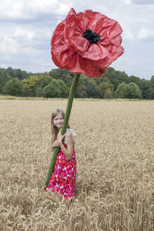 Porträt eines lächelnden Mädchens, das auf einem Feld mit einer übergroßen roten Kunstblume steht - PSTF00250