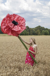 Smiling girl standing on a field with oversized red artificial flower - PSTF00251