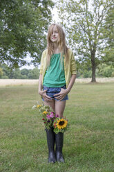 Porträt eines blonden Mädchens, das auf einer Wiese mit Blumensträußen in ihren Gummistiefeln steht - PSTF00252