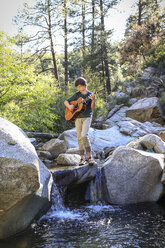 Jugendlicher spielt Gitarre, während er auf einem Felsen in einem Bach in einer natürlichen Landschaft steht - AURF05667