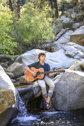 Jugendlicher spielt Gitarre und sitzt auf einem Felsen in einem Bach in einer natürlichen Landschaft - AURF05668