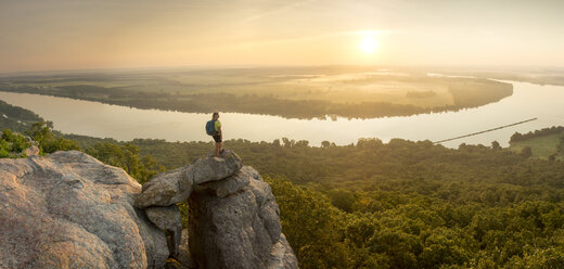 Frau, die auf einem Sandsteinüberhang steht und den Sonnenaufgang vom Gipfel des Petit Jean Mountain über dem Arkansas River Valley beobachtet - AURF05767