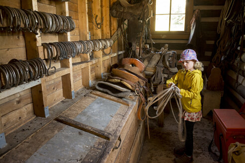 Junges Mädchen wickelt ein Seil in der Sattelkammer auf, während sie sich für einen Ausritt vorbereitet - AURF05770