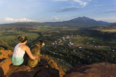 Junge Frau und ihr Hund mit Blick auf einen Berg und eine Stadt bei Sonnenuntergang - AURF05780