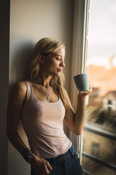 Blonde junge Frau hält Kaffeebecher am Fenster - KKAF01981