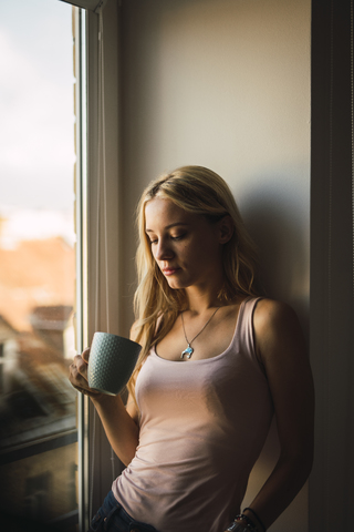 Blonde junge Frau hält Kaffeebecher am Fenster, lizenzfreies Stockfoto