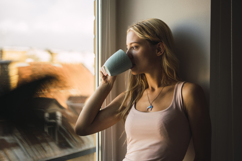 Blond young woman drinking coffee from mug looking out of window stock photo