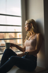 Blond young woman holding coffee mug sitting in windowsill - KKAF01984