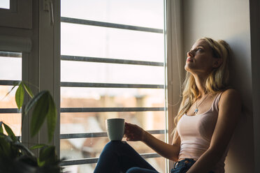 Blond young woman holding coffee mug sitting in windowsill - KKAF01985