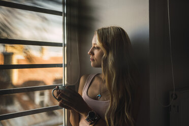 Blond young woman holding coffee mug looking out of window - KKAF01986