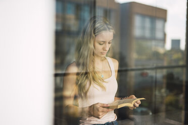 Young woman reading a book behind windowpane - KKAF01989