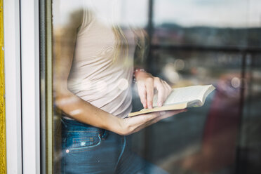 Nahaufnahme einer jungen Frau, die hinter einer Fensterscheibe ein Buch liest - KKAF01992