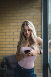 Blonde junge Frau überprüft ihr Handy am Fenster - KKAF01998