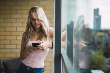 Blond young woman checking cell phone at the window - KKAF01999