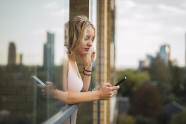 Blond young woman leaning out of window checking cell phone - KKAF02001