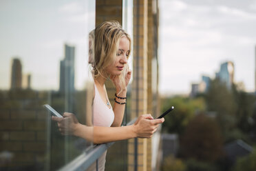 Blonde junge Frau, die sich aus dem Fenster lehnt und ihr Mobiltelefon überprüft - KKAF02001