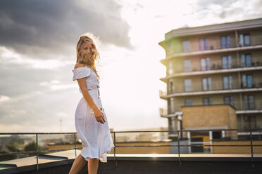 Blonde junge Frau auf Dachterrasse bei Sonnenuntergang in weißem Kleid - KKAF02008