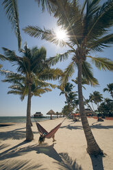 Eine junge Frau lehnt sich in einer Hängematte zwischen Palmen am Strand zurück - AURF05926