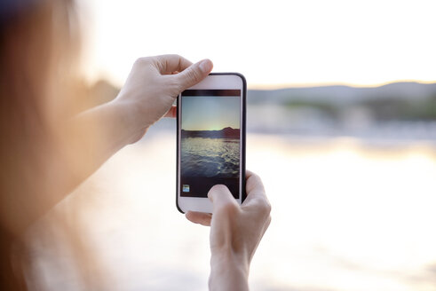 Nahaufnahme einer Frau, die mit ihrem Handy ein Foto vom Meer bei Sonnenuntergang macht - PSIF00105