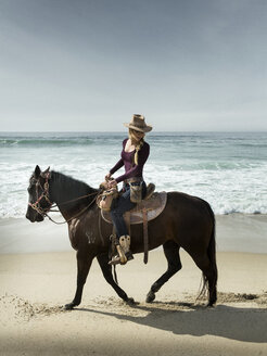 Woman in cowboy hat riding horse on beach - AURF06081