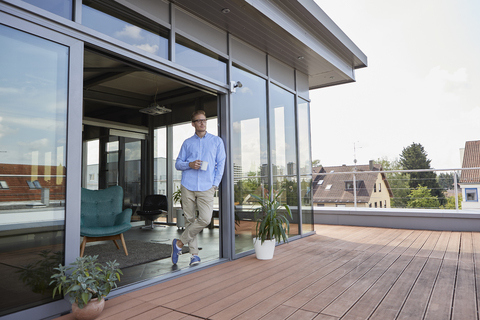 Young man with cup of coffee standing at balcony door stock photo