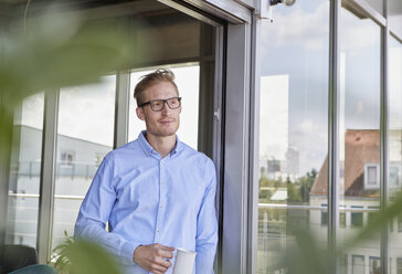 Young man with cup of coffee standing at balcony door - RBF06801