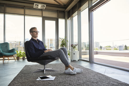 Lächelnder junger Mann im Sessel am Fenster zu Hause sitzend - RBF06807