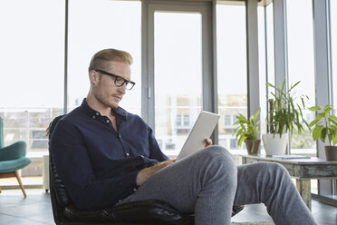 Junger Mann sitzt im Sessel am Fenster zu Hause und benutzt ein Tablet - RBF06814