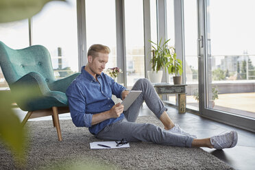 Junger Mann sitzt zu Hause auf einem Teppich und benutzt ein Tablet - RBF06817