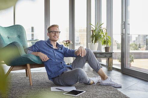 Smiling young man sitting on carpet with tablet and notepads at home - RBF06819