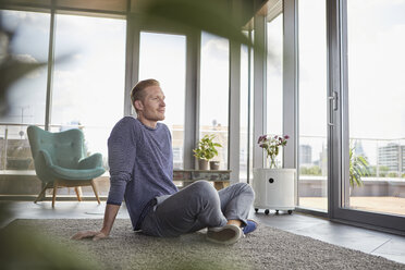 Young man sitting on carpet at home looking out of window - RBF06820