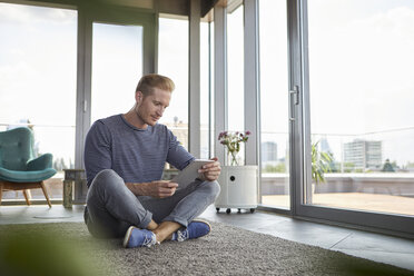 Junger Mann sitzt zu Hause auf einem Teppich und benutzt ein Tablet - RBF06821