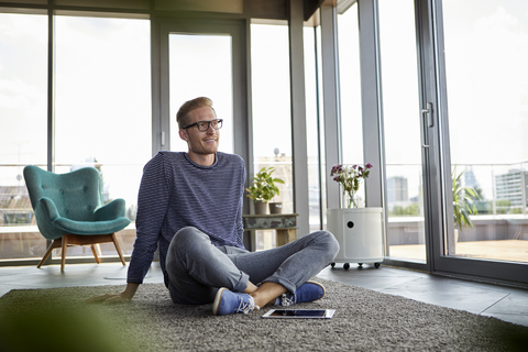 Lächelnder junger Mann sitzt mit Tablet zu Hause auf dem Teppich, lizenzfreies Stockfoto