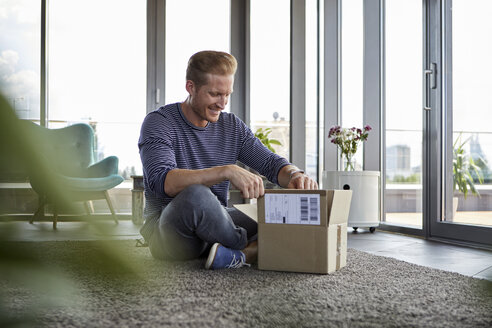 Smiling young man sitting on carpet at home unpacking parcel - RBF06826