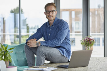 Porträt eines lächelnden jungen Mannes, der mit einem Laptop auf einem Tisch sitzt - RBF06837