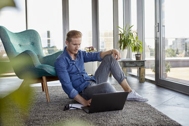 Junger Mann sitzt zu Hause auf einem Teppich und benutzt einen Laptop - RBF06845