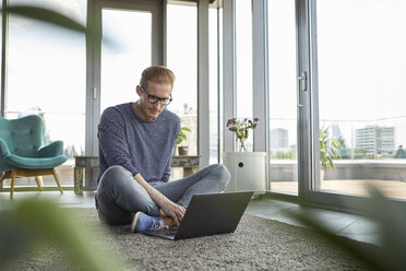 Junger Mann sitzt zu Hause auf einem Teppich und benutzt einen Laptop - RBF06846