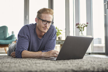 Young man lying on carpet at home using laptop - RBF06847