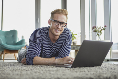 Porträt eines lächelnden jungen Mannes, der zu Hause auf dem Teppich liegt und einen Laptop benutzt, lizenzfreies Stockfoto